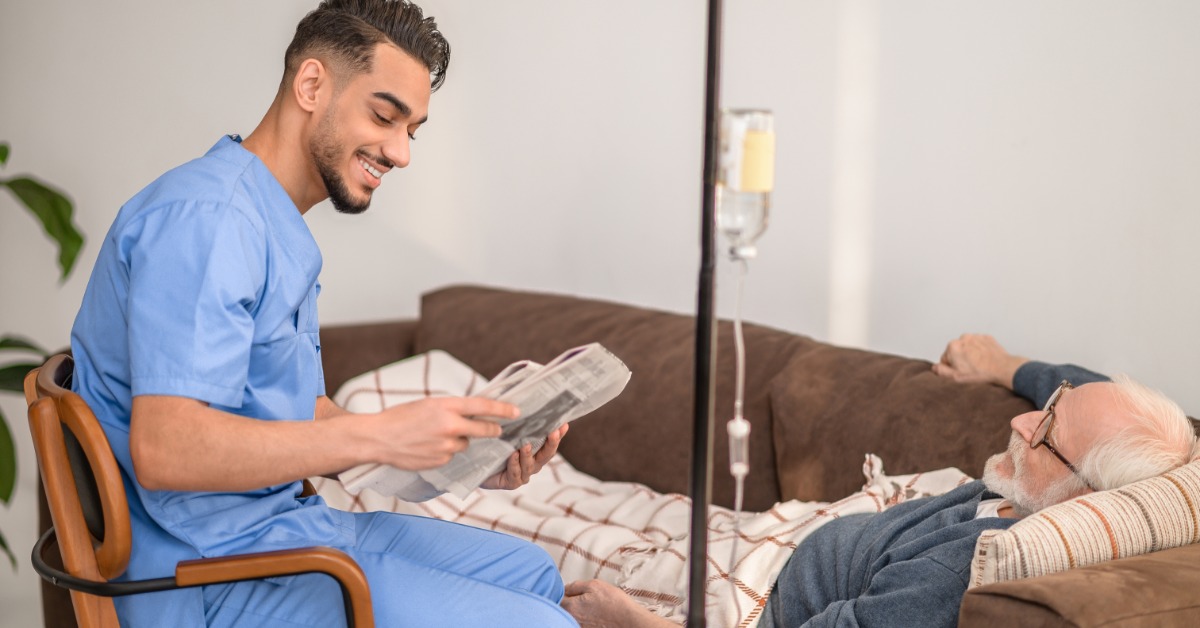 A friendly male nurse in blue scrubs smiles while talking to an older man lying on a brown couch with a blanket.
