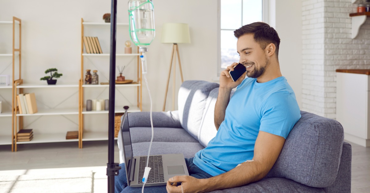 A man wearing a blue shirt sits on a couch at home, talking on the phone while getting a mobile IV treatment.
