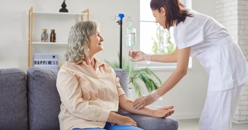 A young female medical nurse helps administer an IV to a woman sitting on a couch at home.