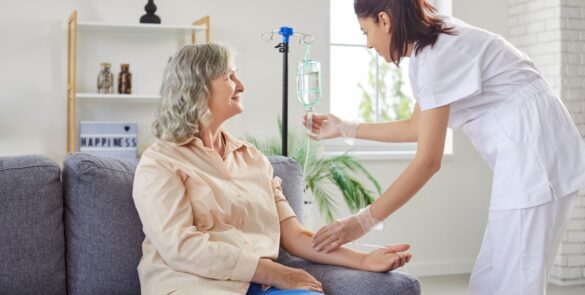 A young female medical nurse helps administer an IV to a woman sitting on a couch at home.
