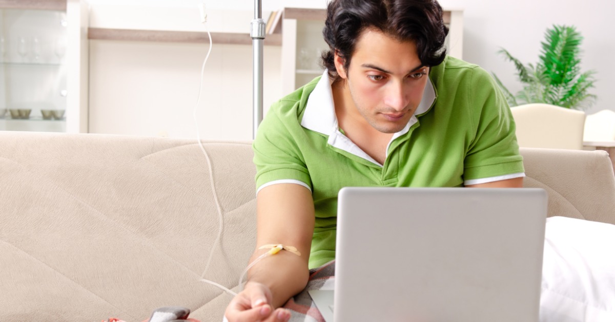A young man sitting at home in front of a laptop on the couch, connected to an IV drip behind the couch.