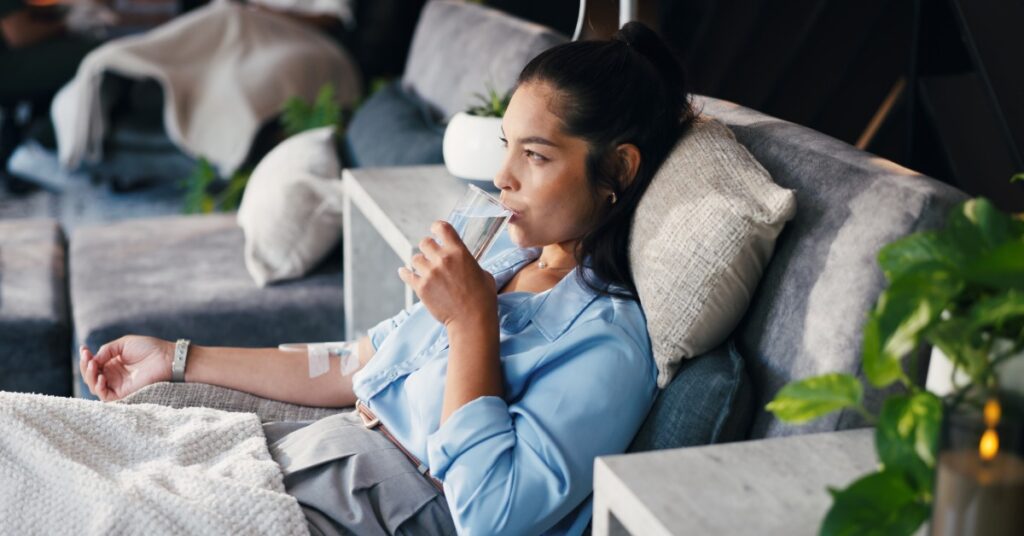 A woman lying on a couch, drinking a glass of water in one hand while her other arm in lying flat, connected to an IV.