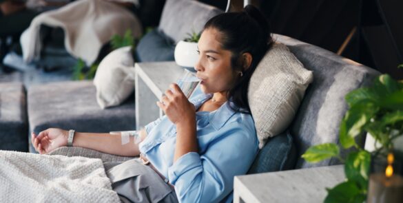 A woman lying on a couch, drinking a glass of water in one hand while her other arm in lying flat, connected to an IV.