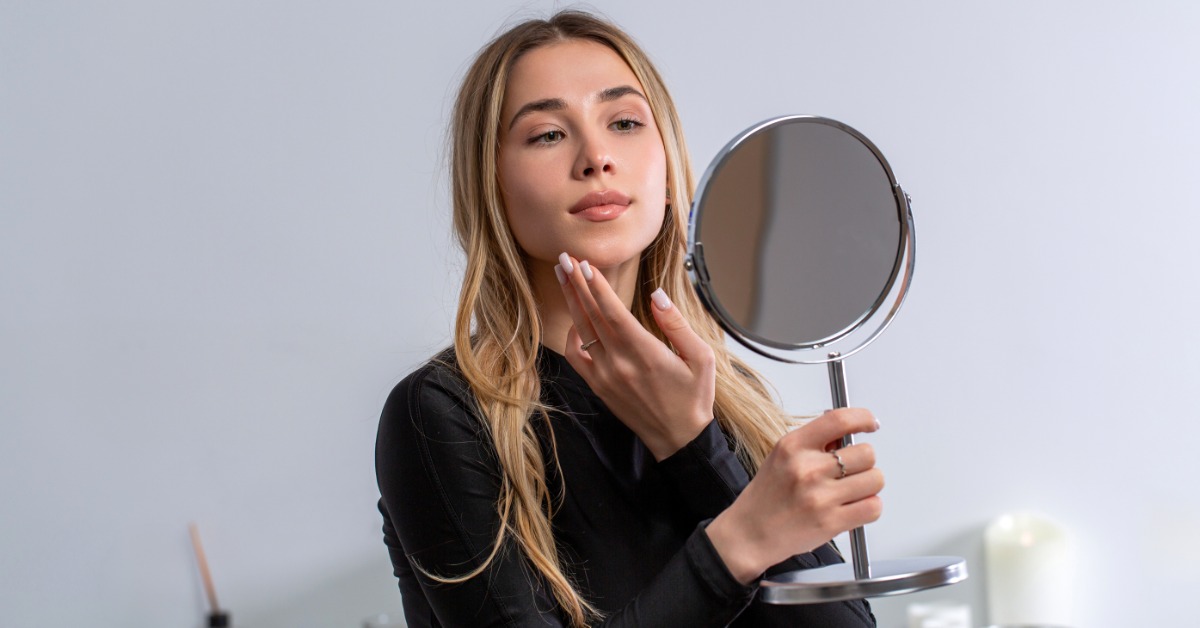 A young woman holding a hand mirror up in front of her and examining her skina nd face in the reflection.