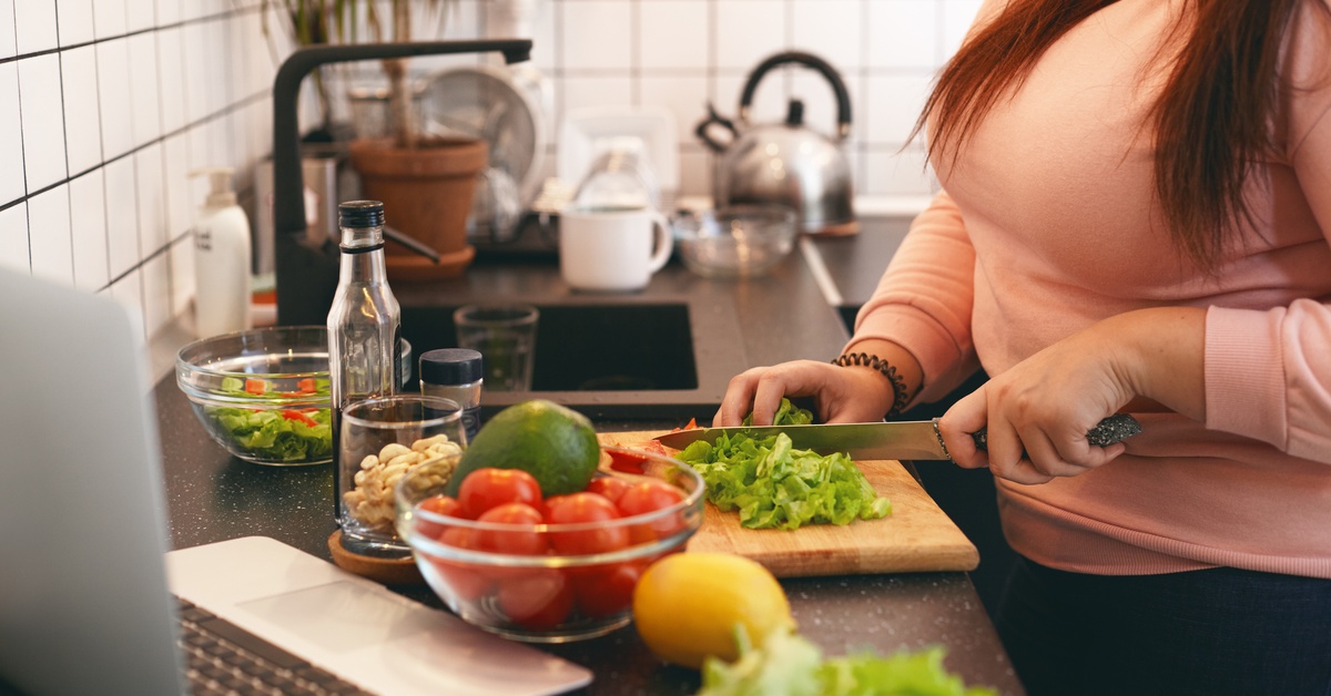 a person cutting fresh vegetables on a kitchen counter beside a laptop, bowl, cutting board, and utensils.