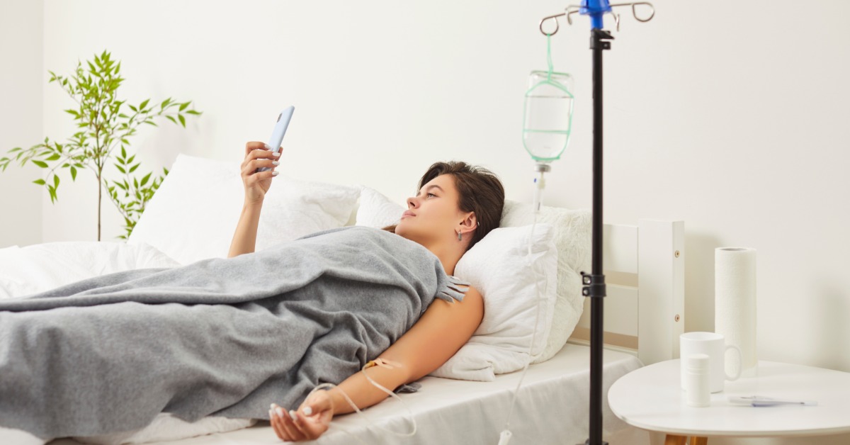 a woman lying in bed under a gray blanket while holding a smartphone beside an IV stand and a small table.