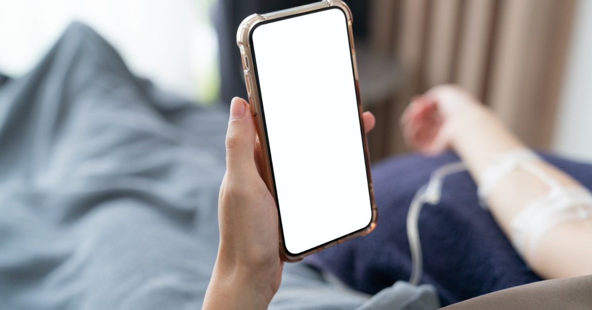 a hand holding a smartphone with a blank white screen while an IV line is taped to an arm near gray bedding.