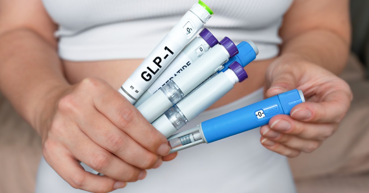 Close-up of hands holding multiple medication injection pens, including a white pen prominently labeled GLP-1 in black.