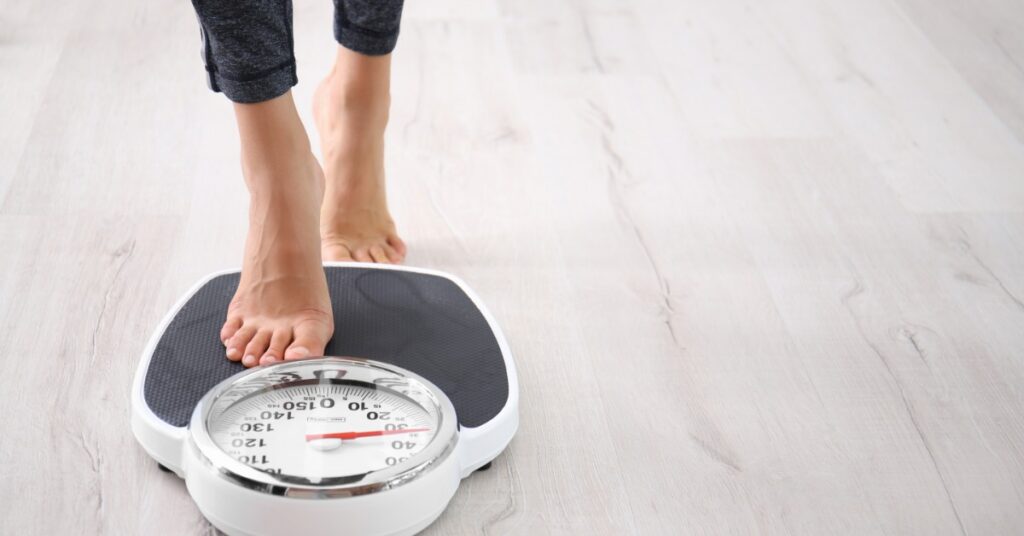 Person steps barefoot onto a white analog bathroom scale with a black platform and round numbered dial on a wood floor.