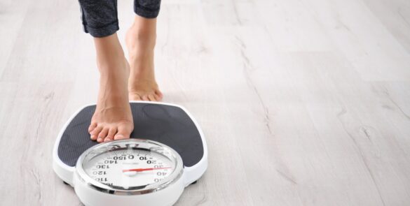 Person steps barefoot onto a white analog bathroom scale with a black platform and round numbered dial on a wood floor.