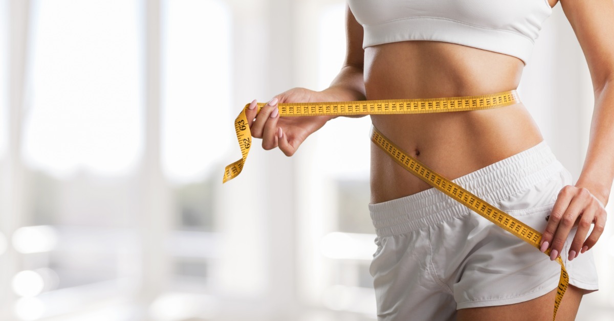 Cropped view of a person in white workout clothes holding a yellow measuring tape around their waist and hips.