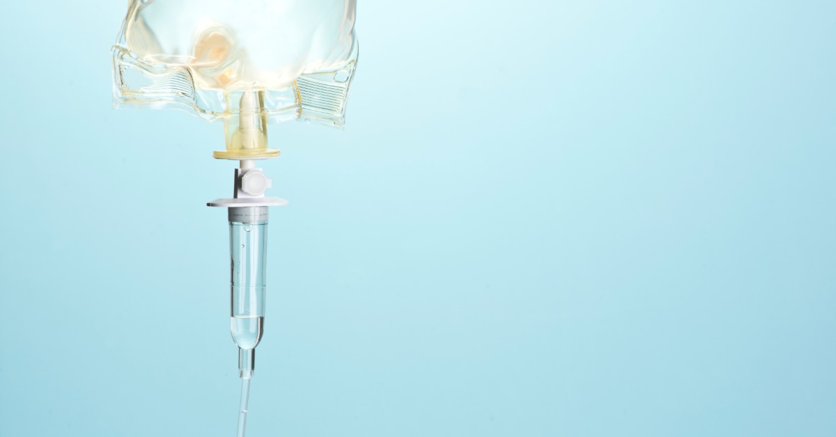 A clear IV bag hanging from a metal stand with tubing and a blurred indoor room with equipment in the background.