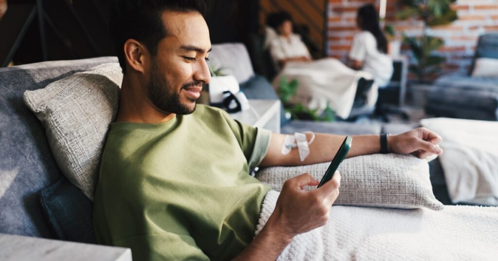 A man sitting in a lounge chair using a smartphone while an IV bag hangs beside him on a metal stand.