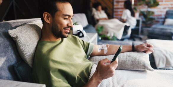A man sitting in a lounge chair using a smartphone while an IV bag hangs beside him on a metal stand.