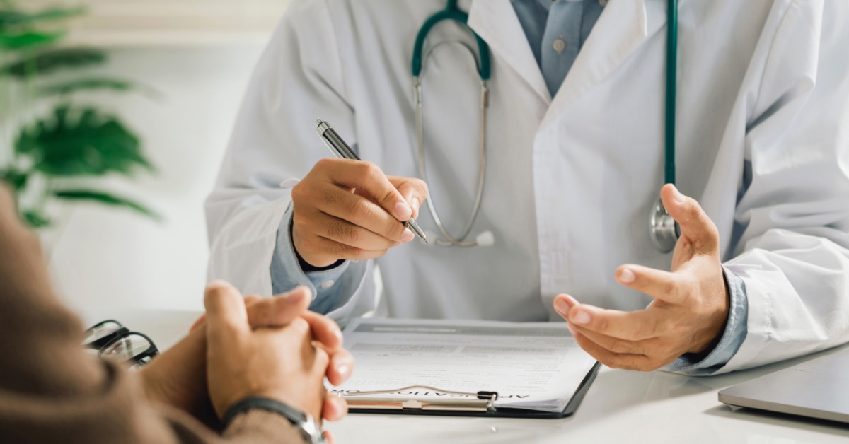 A person in a white coat writing on paperwork across a desk from a seated man in a medical office during a consultation.
